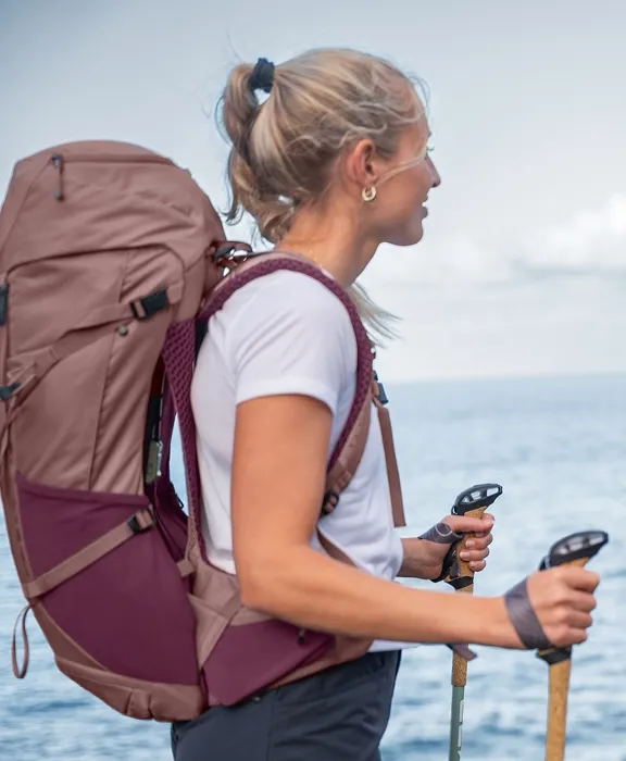 Eine Frau von der Seite mit einem weinroten Deuter-Trekking Rucksack und Wanderstöcken vor einem Meerblick.