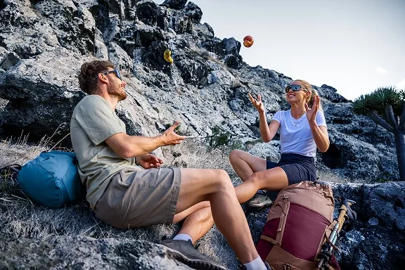 Zwei Wanderer machen lachend Pause auf Felsen; neben ihnen zwei Wanderrucksäcke.